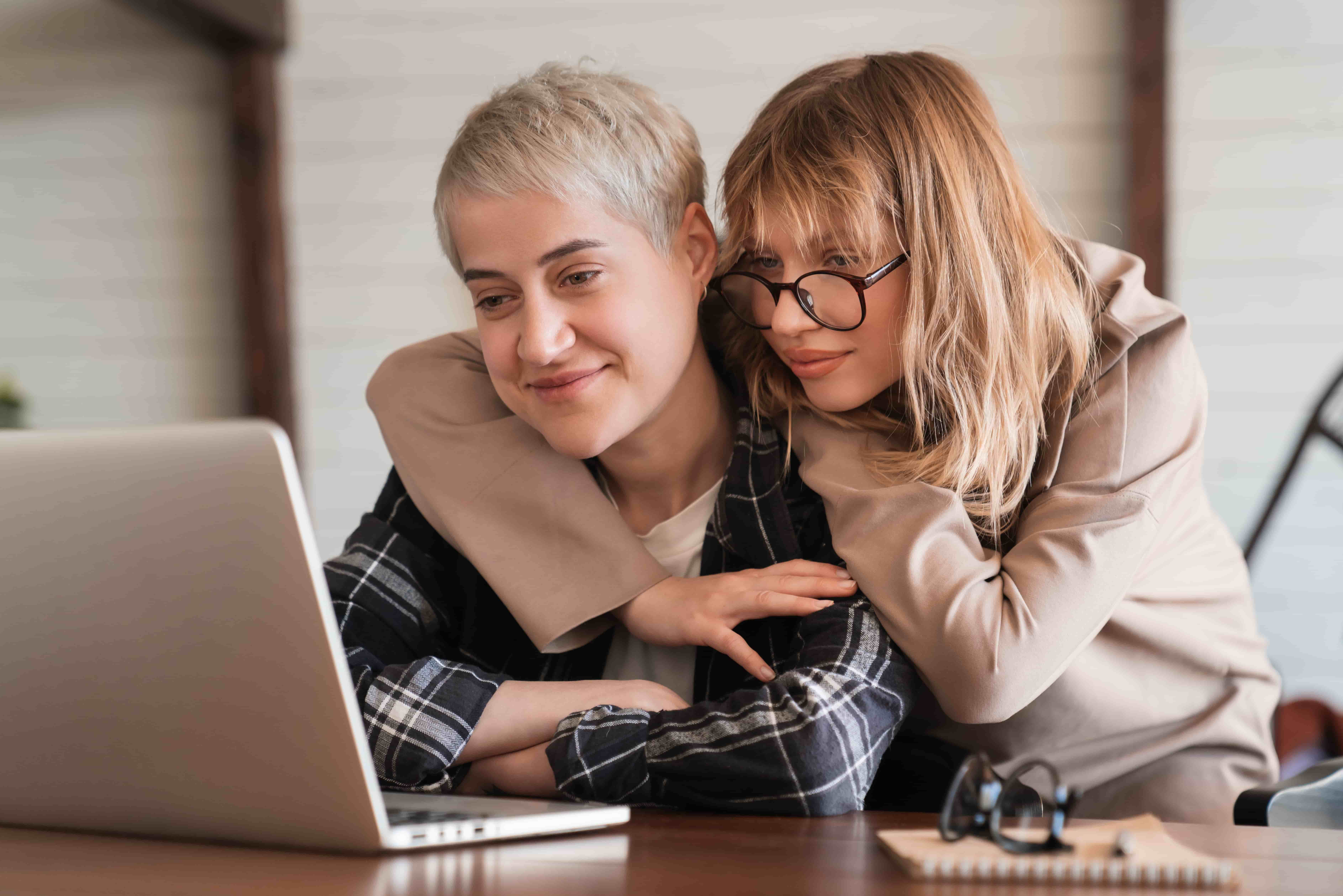 Two female LGBT lesbian couple best girlfriends students hugging embracing watching movie webinar on laptop studying e-learning together, doing paperwork, paying bills online in home office